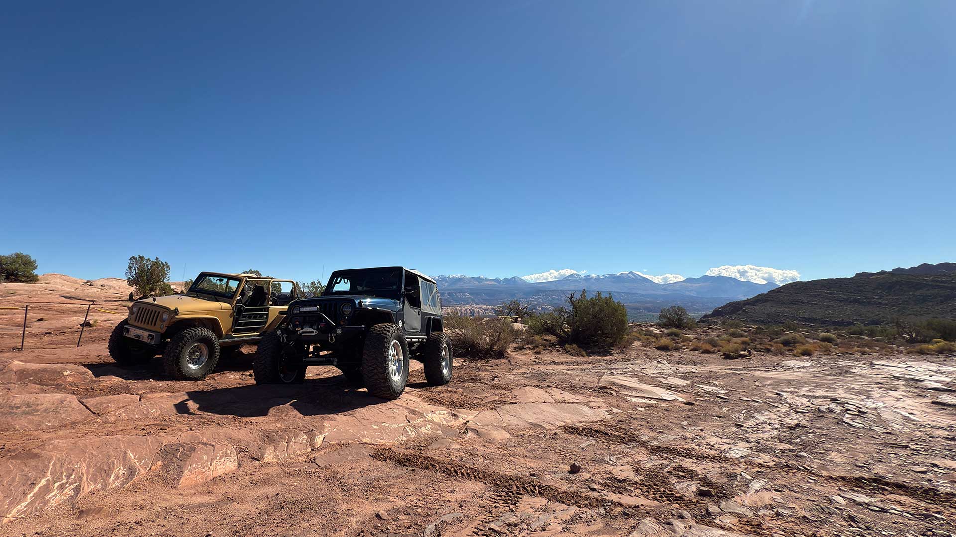 Two In Too Deep rigs, a yellow Jeep and a blue jeep, posing at Moab with snow-capped mountains in the distance.
